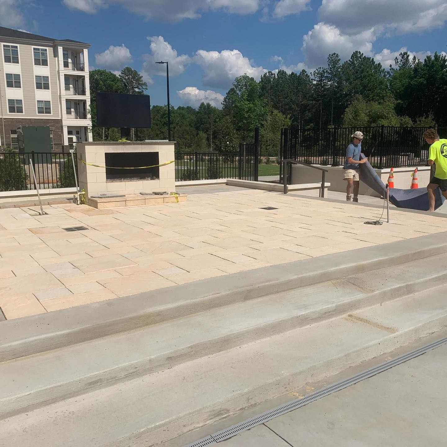 Workers install landscape fabric on a patio with an outdoor fireplace near a building with a blue sky.