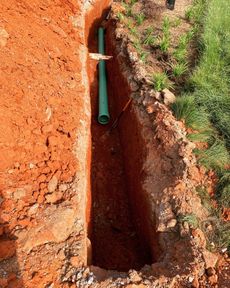 Trench dug in red clay soil with a green pipe in it, next to grass.
