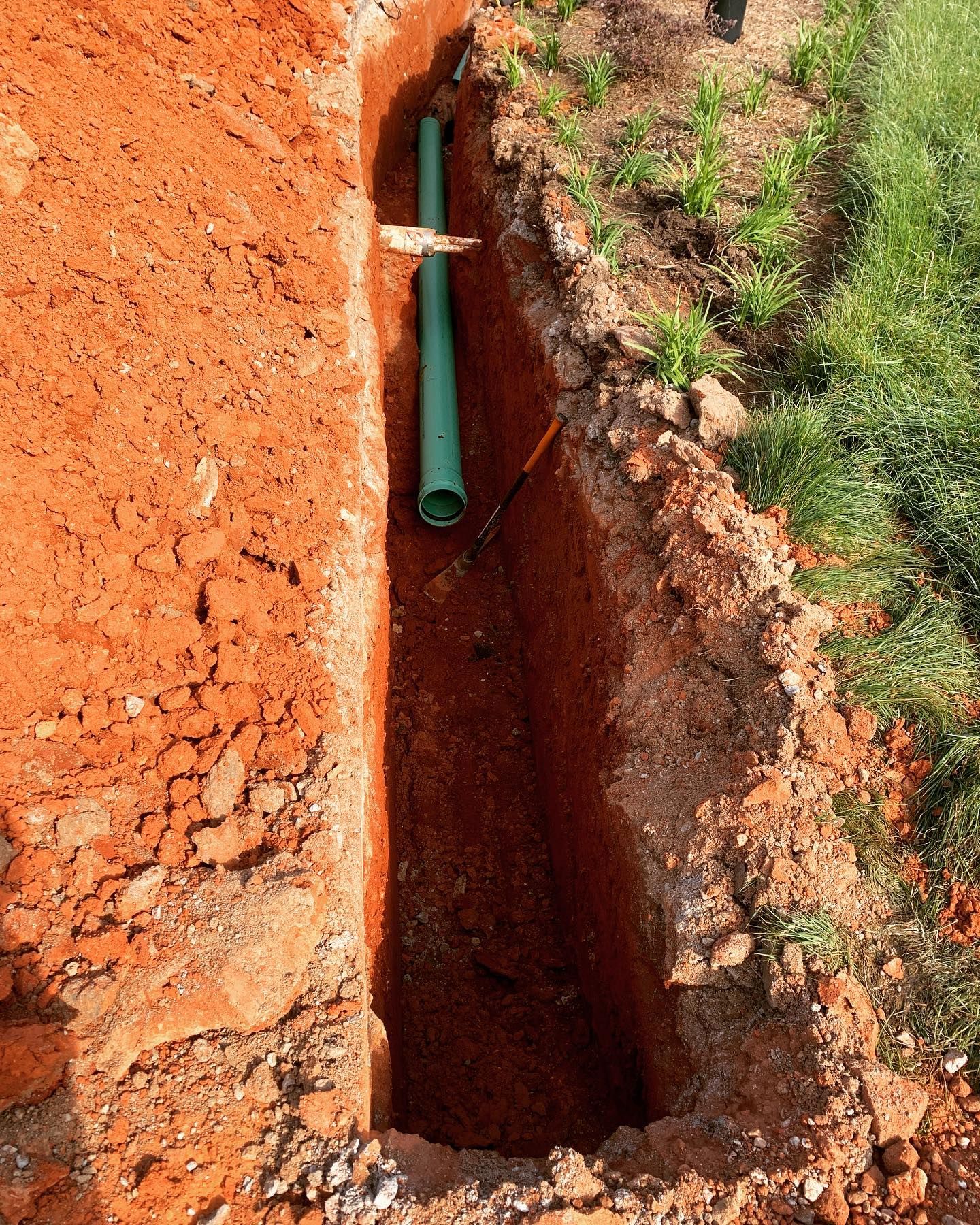 A trench dug in red soil with green pipe inside, near grass.