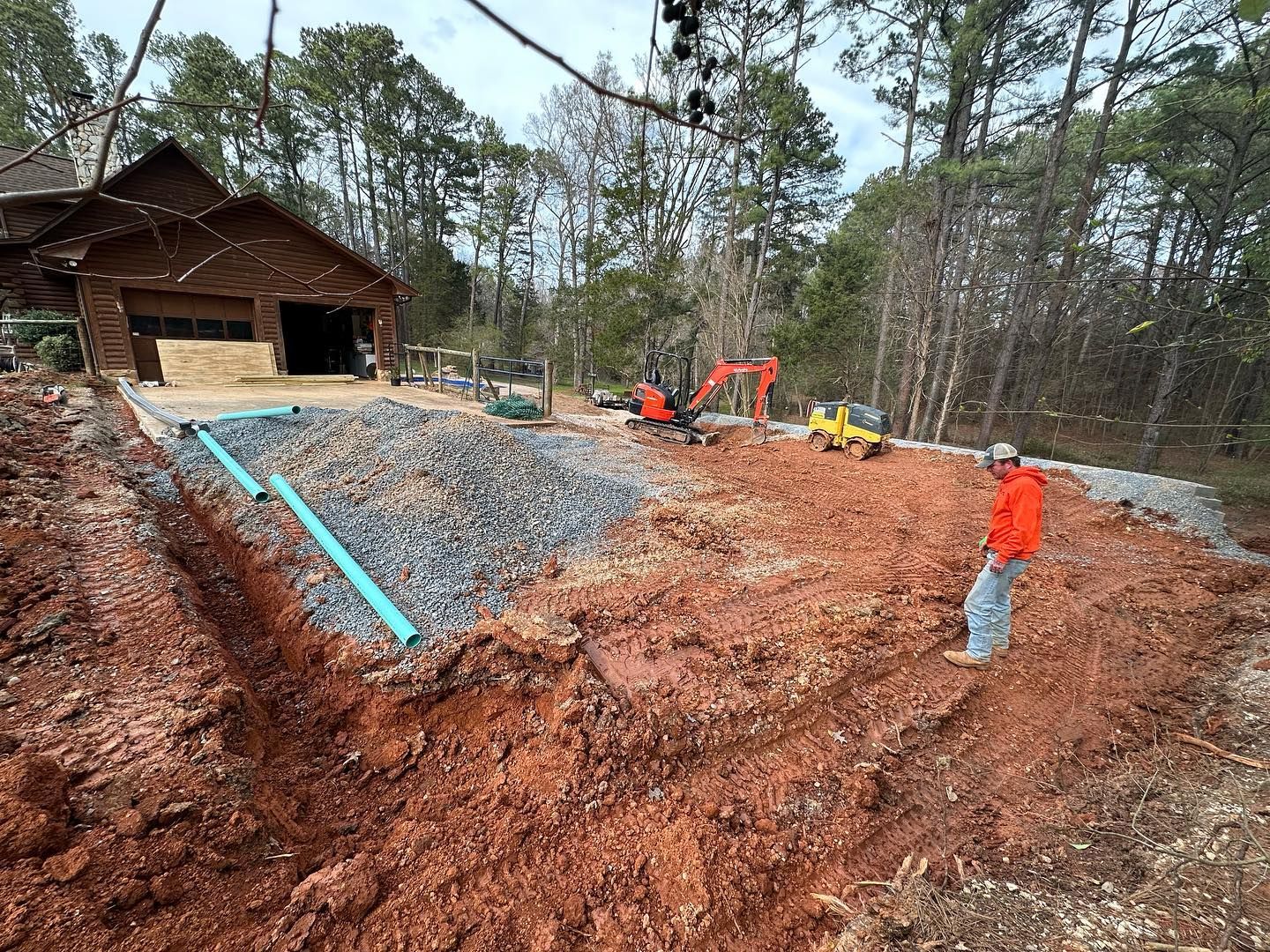 Construction site with a person, machinery, dirt, gravel, and a log cabin in the background.