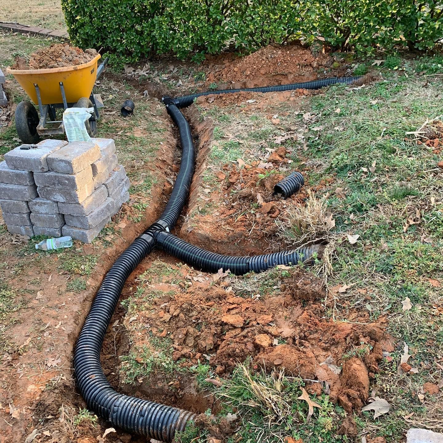 Drainage pipes in a trench, garden setting. Wheelbarrow, bricks, and bushes are visible.
