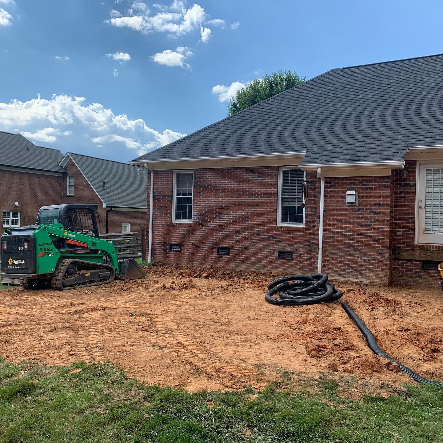 A small skid steer compact track loader digs in a dirt backyard next to a brick house.
