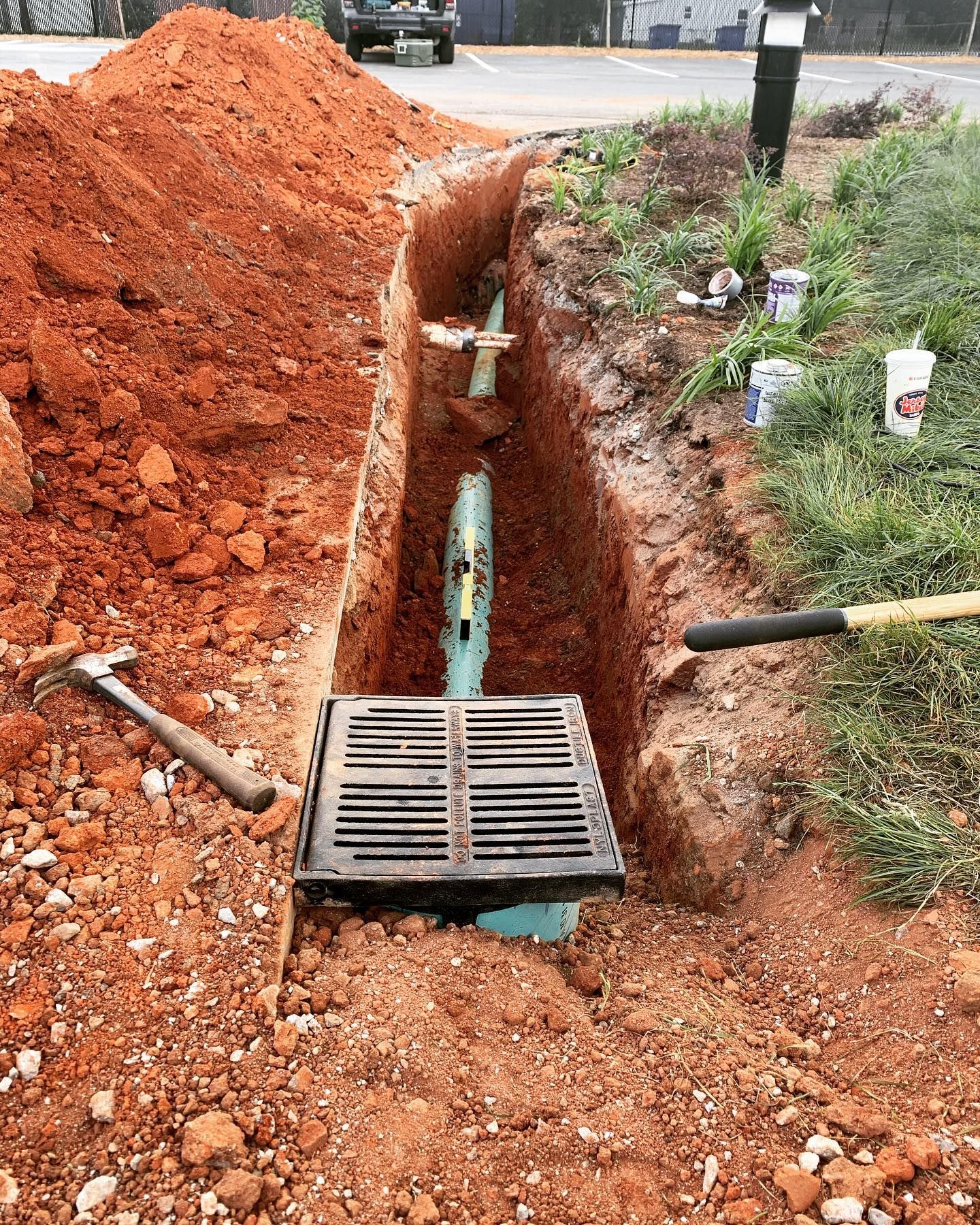 Trench with drain, pipes, and tools in reddish-brown dirt, near grass and parking lot.