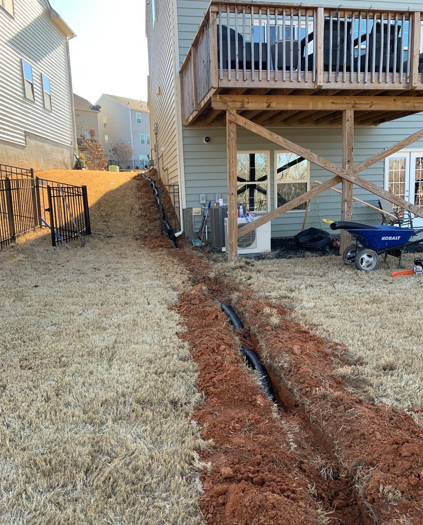 Trench dug in a grassy yard with black pipes; house with deck in the background.