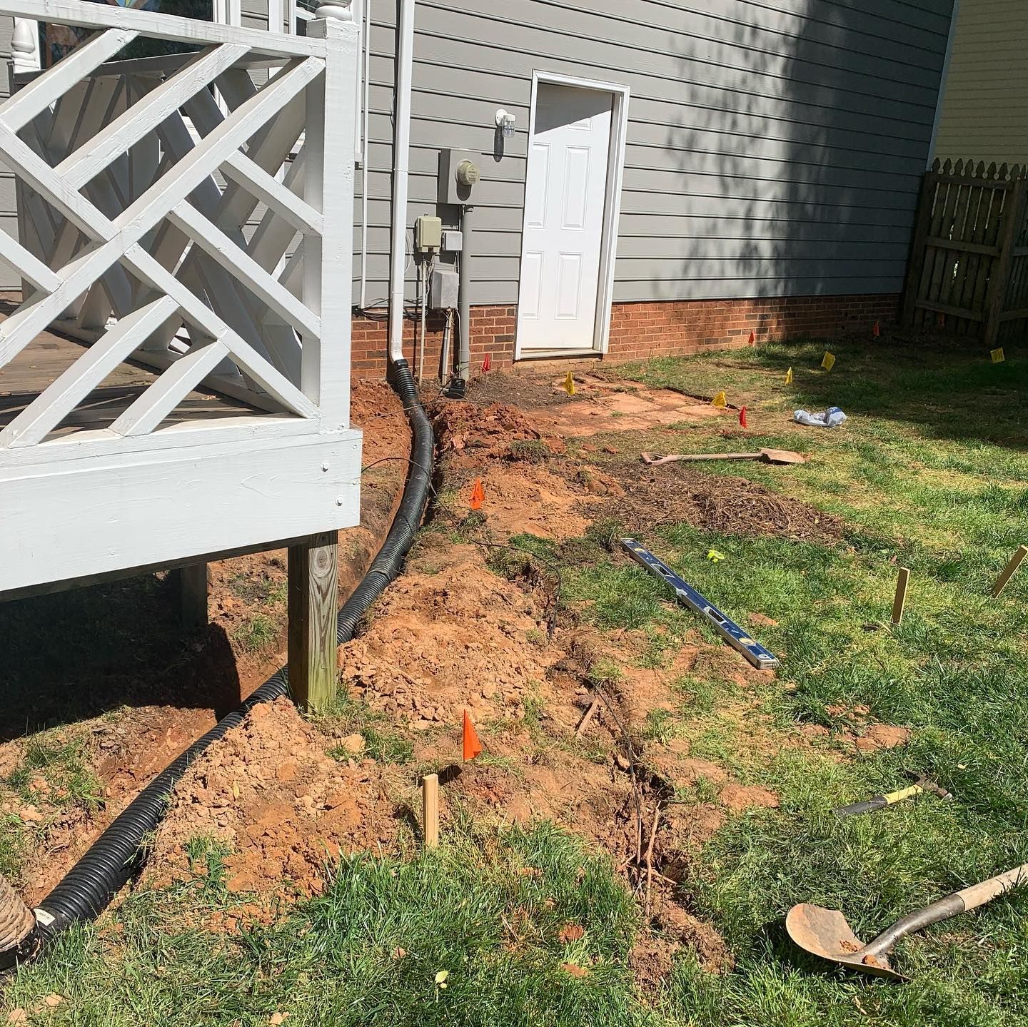 Drainage project near a house. Black pipe runs along a dirt trench, with orange markers.