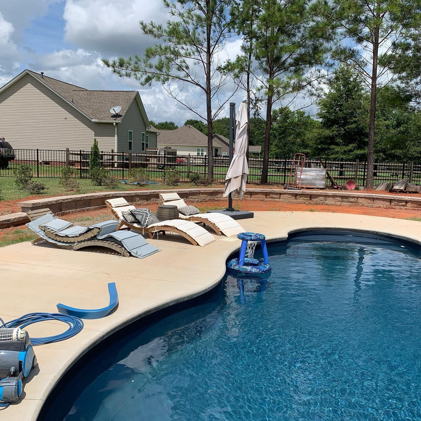 A backyard pool with lounge chairs and a blue pool cleaner, surrounded by a concrete patio and fence.