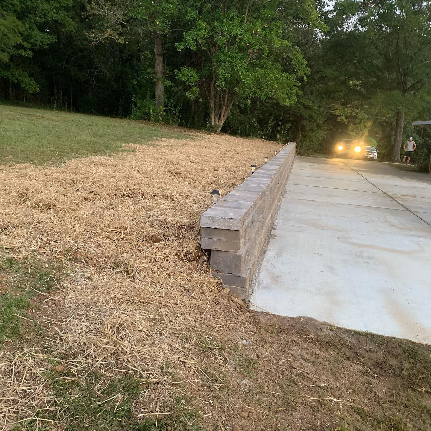 Concrete block retaining wall next to a driveway and dead grass. Car lights in the distance.