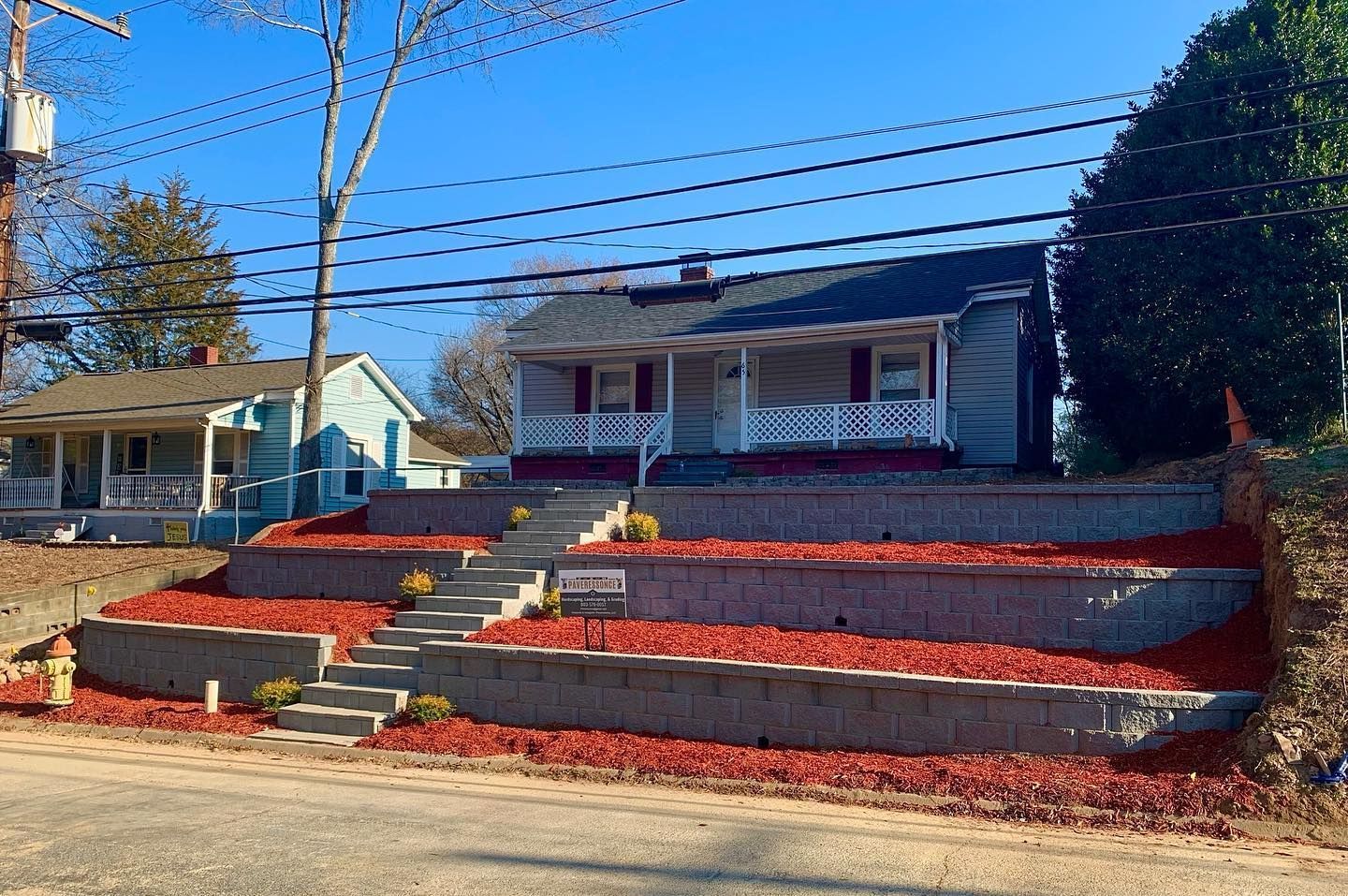 A small, light-colored house on a raised lot with steps and red mulch. A blue house is to the left.