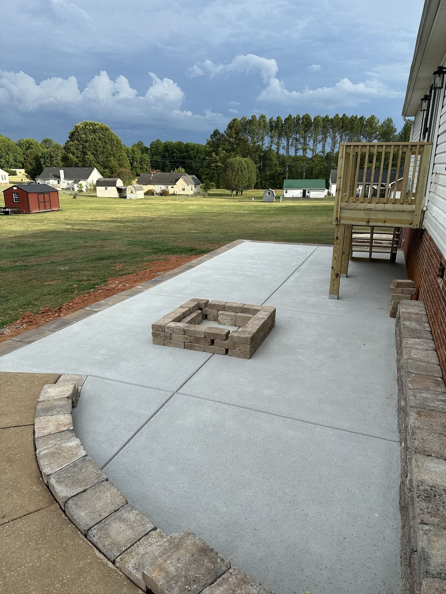 Concrete patio with a brick fire pit and deck, in a grassy backyard.