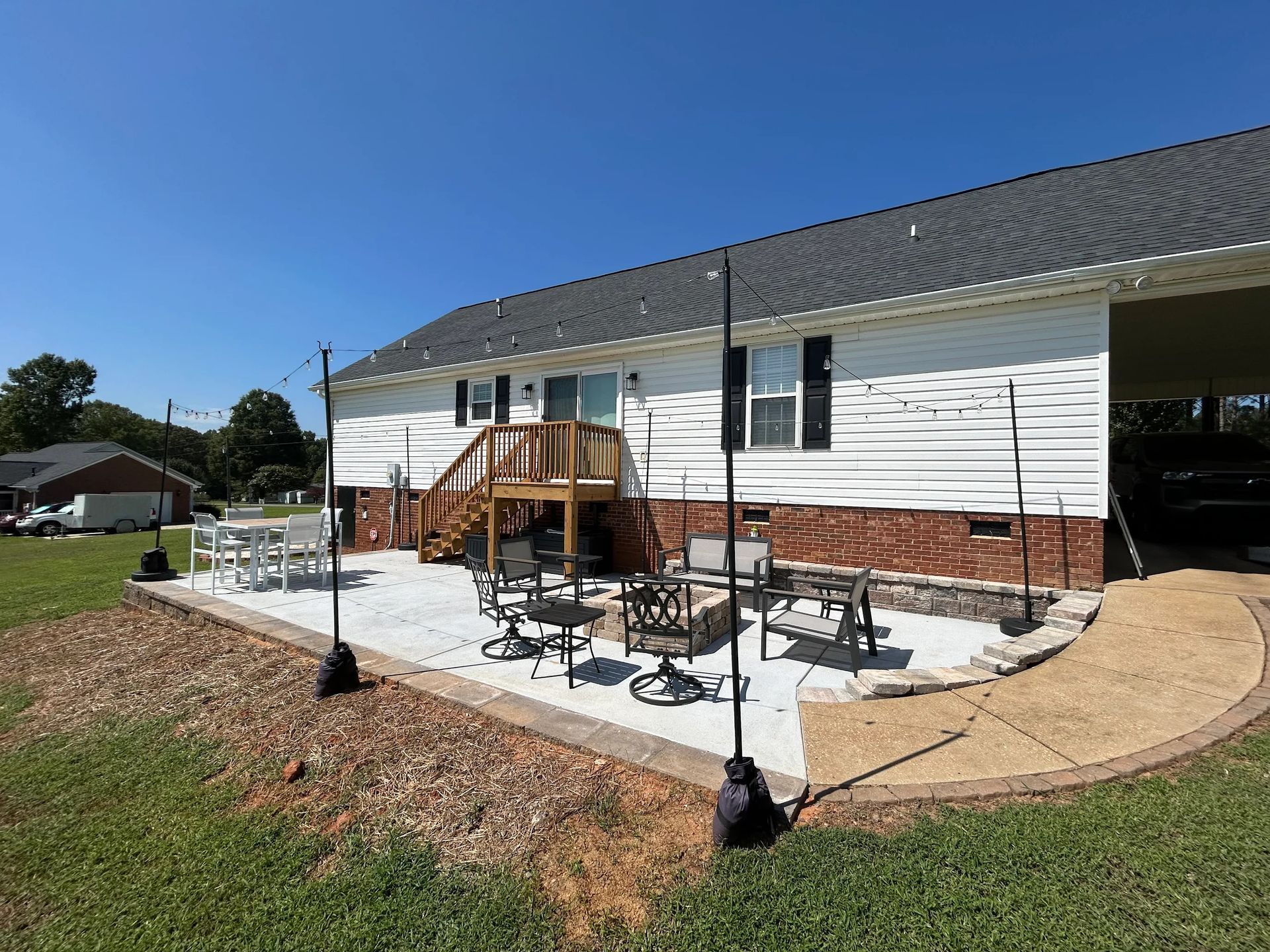Backyard patio with seating, fire pit, and string lights, adjacent to a white house with brick accents and a clear blue sky.