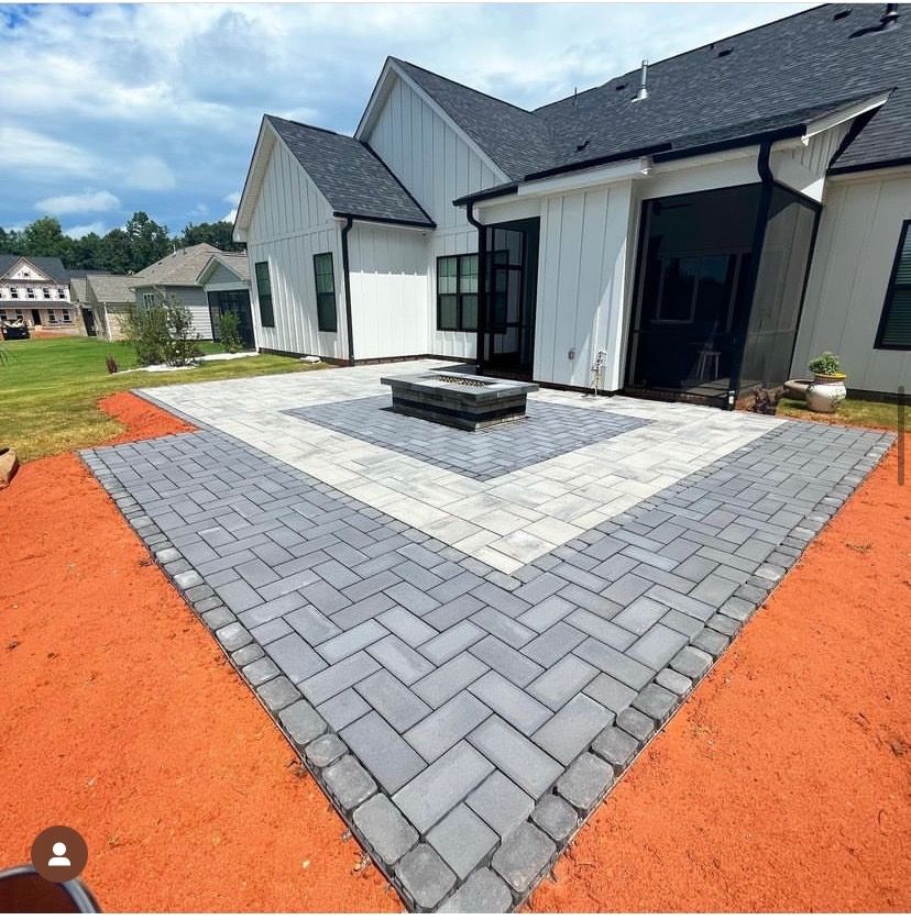 Patio with gray and white pavers, fire pit, next to a white house with black trim, in a yard.