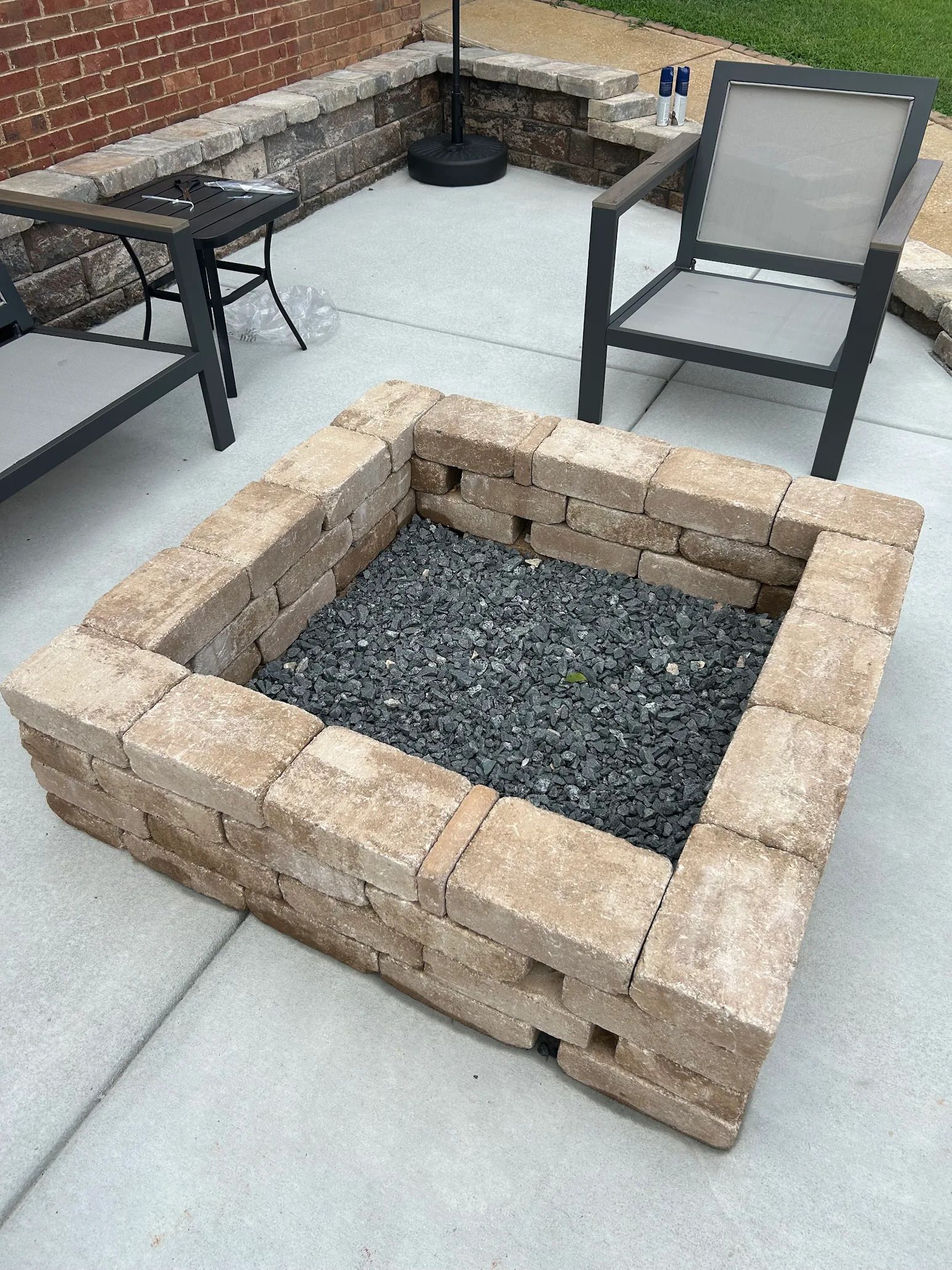 Square stone fire pit filled with dark gray gravel on a patio, with outdoor furniture in the background.