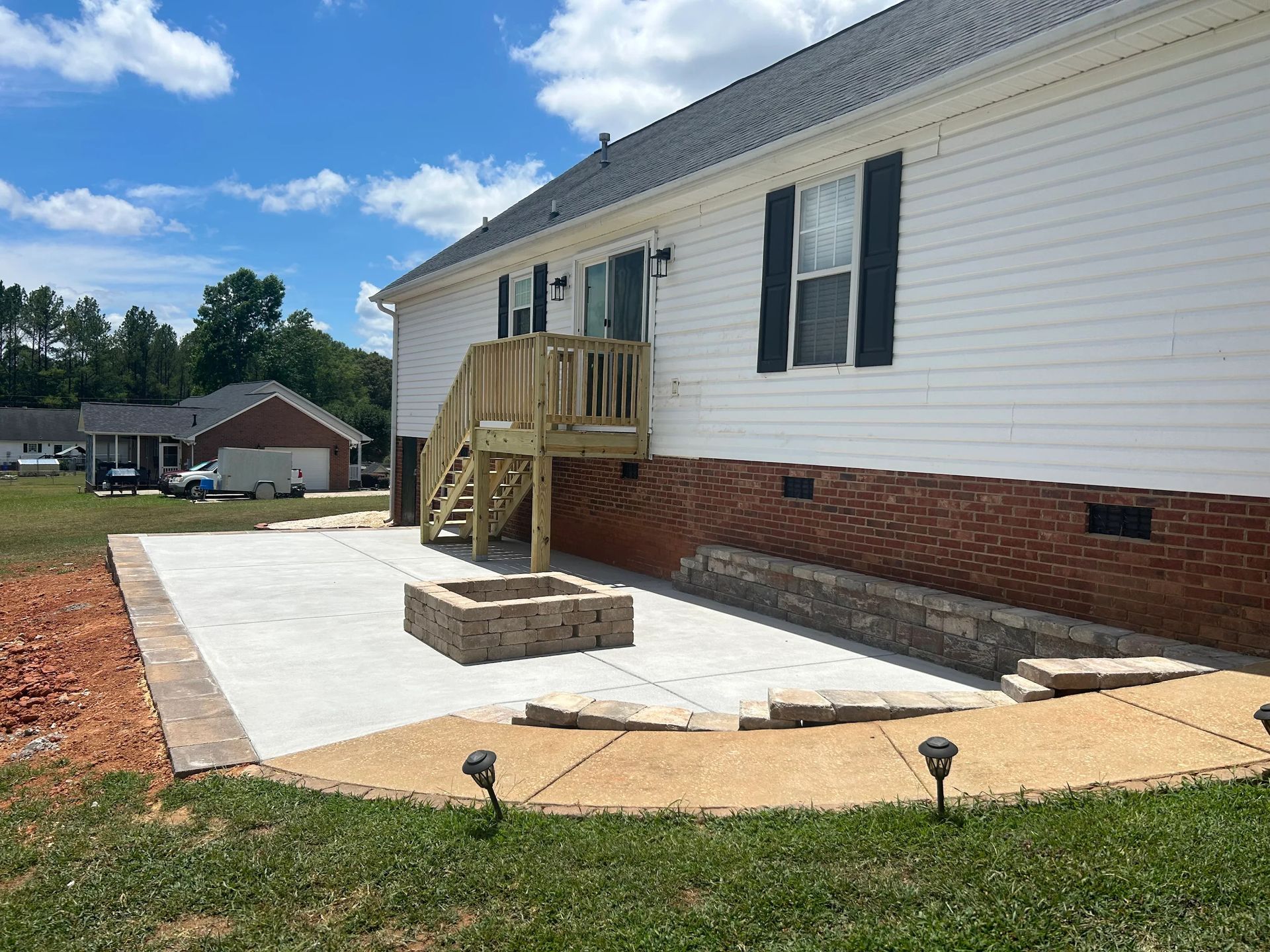Backyard patio with concrete and brickwork, wooden deck stairs, and a fire pit. Sunny day.