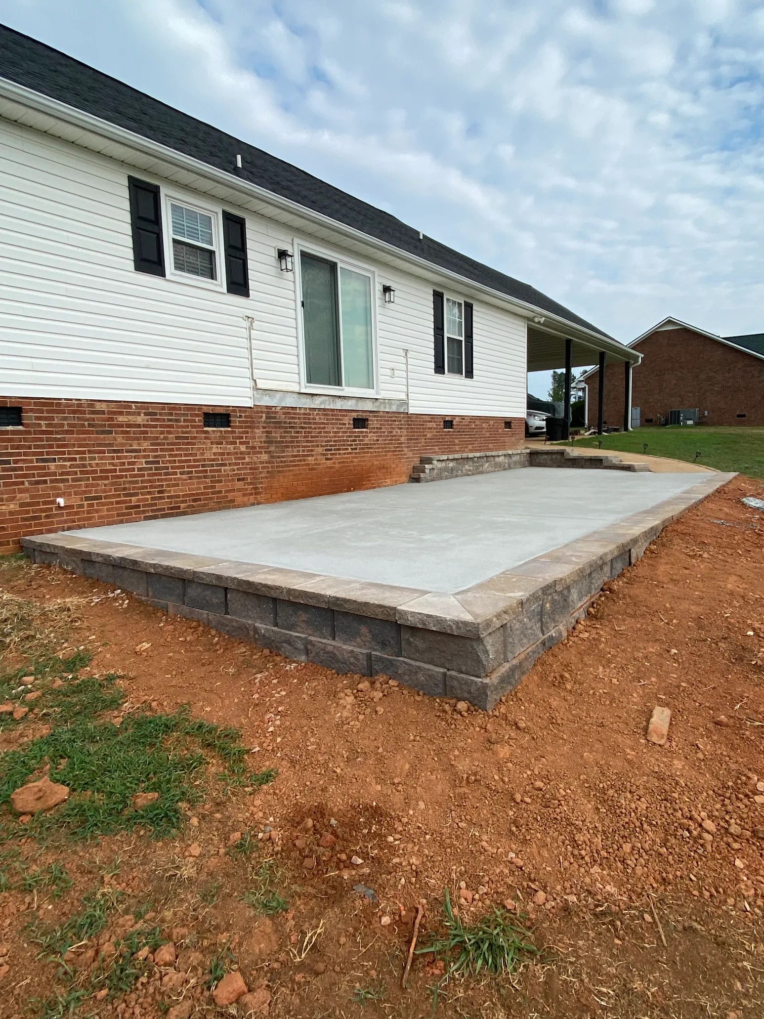 A newly constructed patio with retaining walls next to a white house with a red brick foundation.