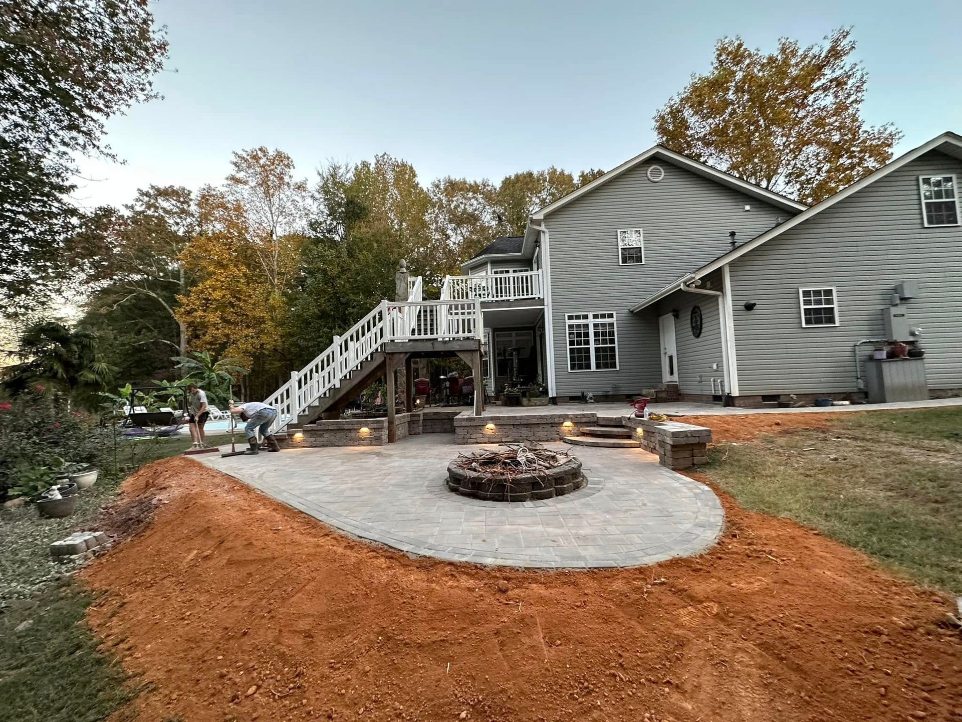 Backyard with a paver patio, fire pit, deck, and house. A person works on the patio.