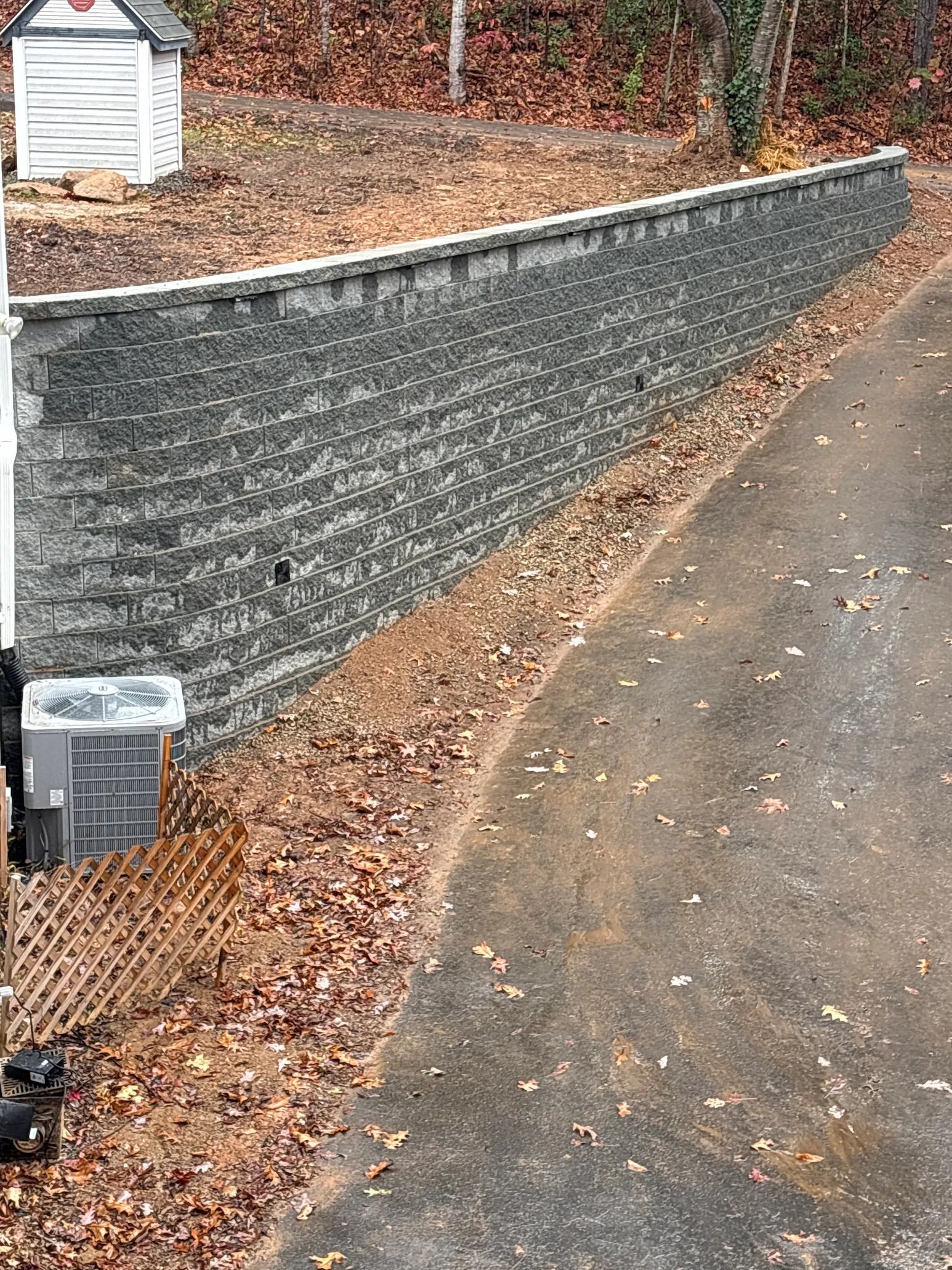 Gray retaining wall next to a road, covered in fallen leaves. Small shed in the background.