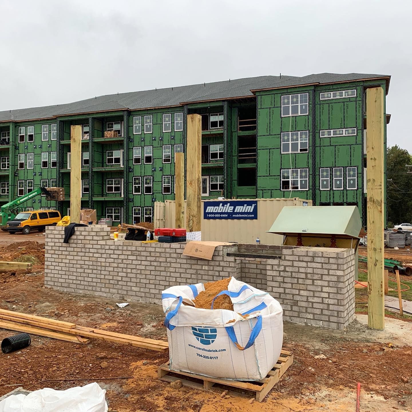 Construction site: multi-story building with green siding, brick wall in progress, large bag of dirt on wooden pallet.