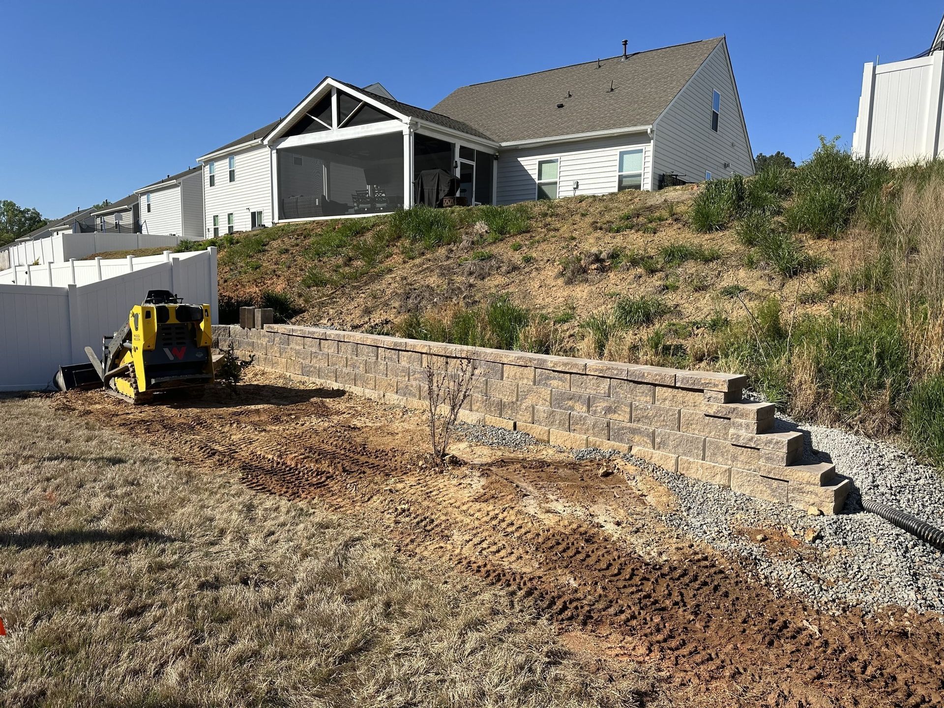 Construction site with retaining wall and mini excavator near a two-story house on a hill.
