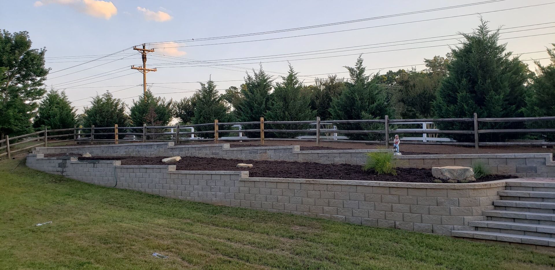 A flock of birds flies over a tiered landscaping of stone walls, trees, a fence, and green grass.