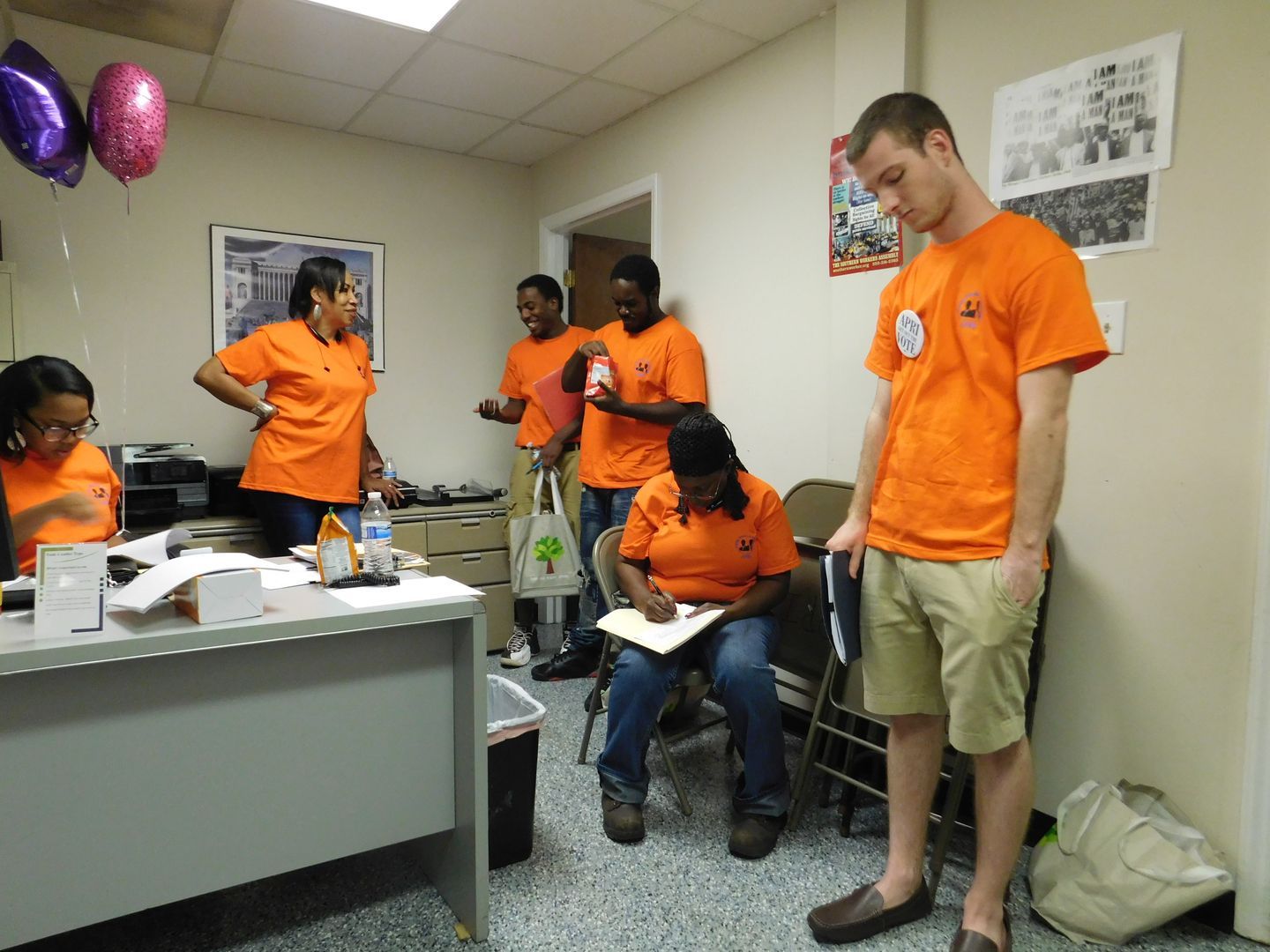 A group of Get Out The Vote volunteers in orange shirts are standing in a room