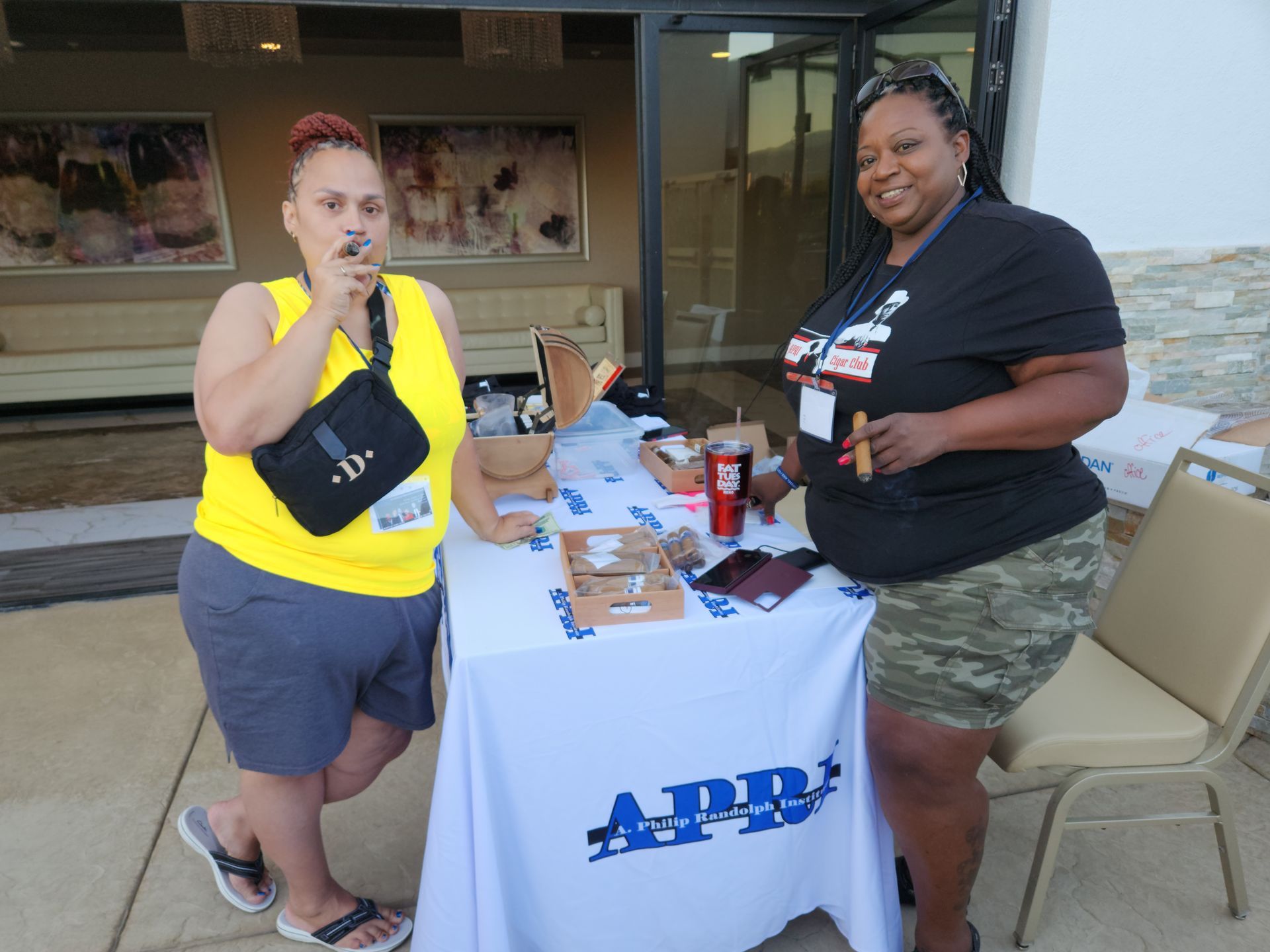 Two women smoking cigars, standing next to a table that says APRI on it