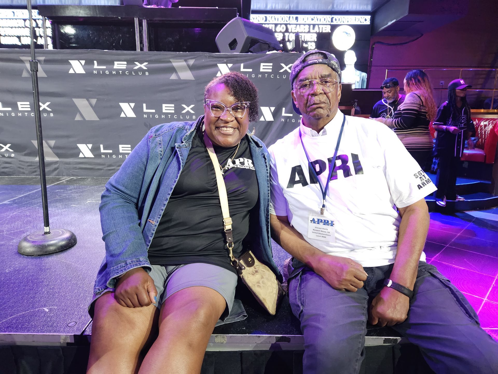 A man with an APRI shirt and a woman are sitting next to each other on a stage.