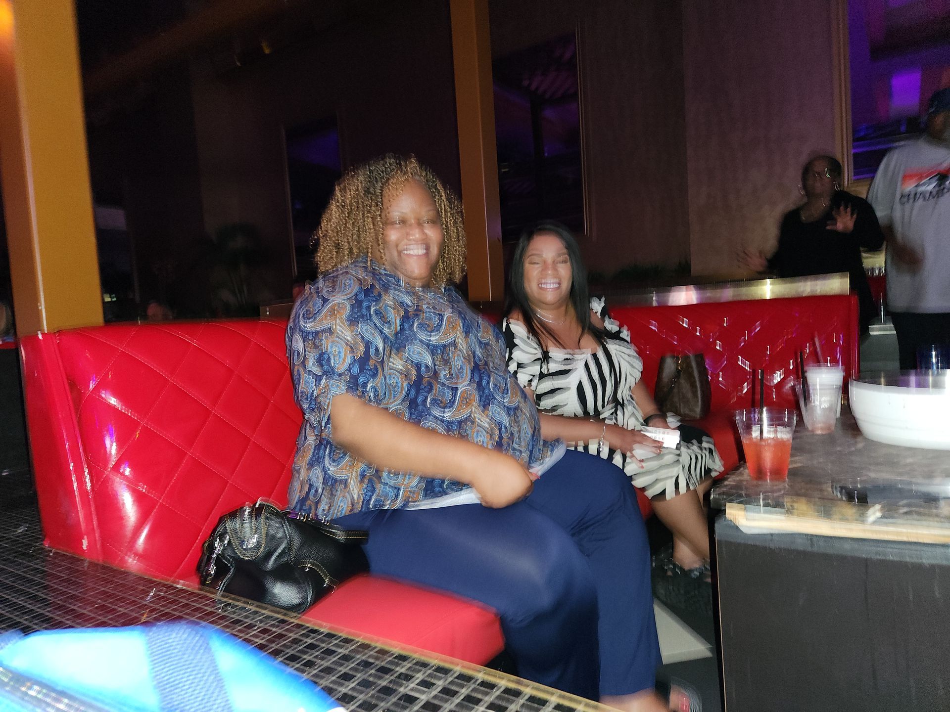 Two women are sitting on a red couch and smiling  at an APRI party