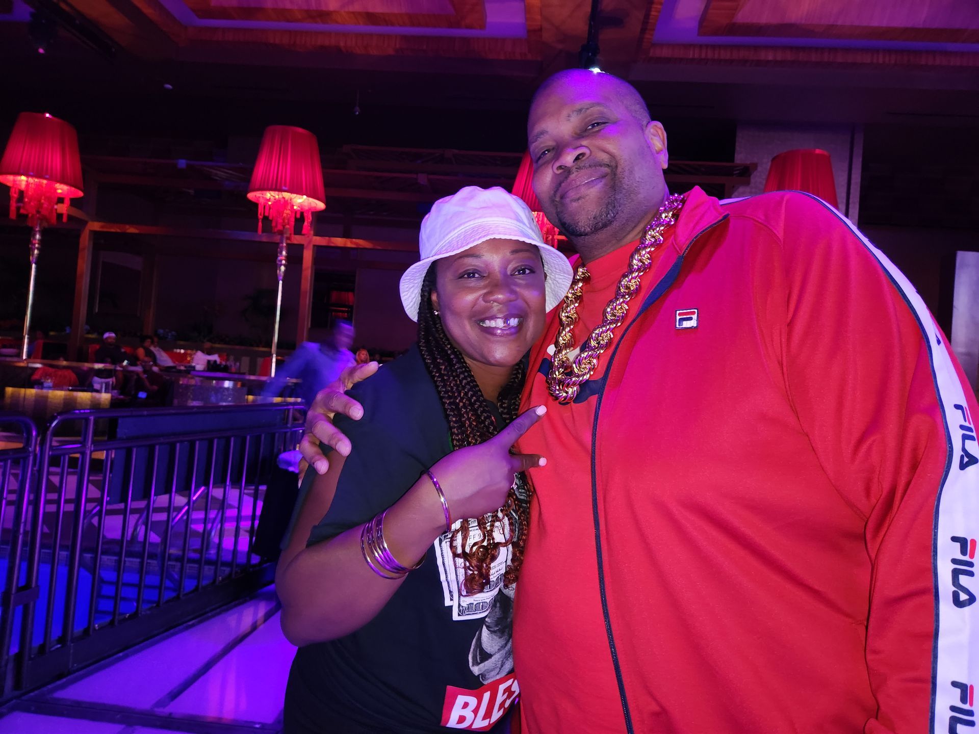 Danny in a red Fila jacket and fat, gold, rope-chain poses with a woman at an APRI 