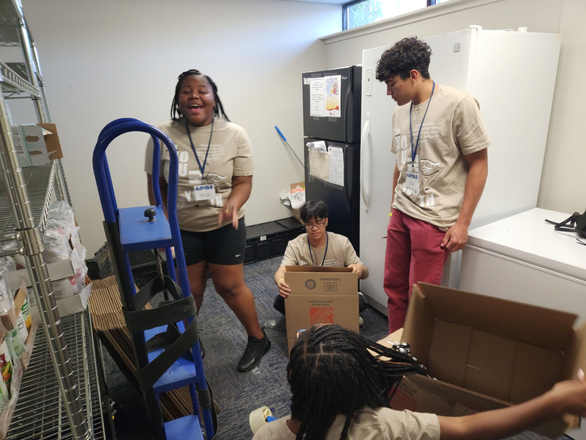 A group of APRI Youth volunteers standing in a room with boxes and a refrigerator.