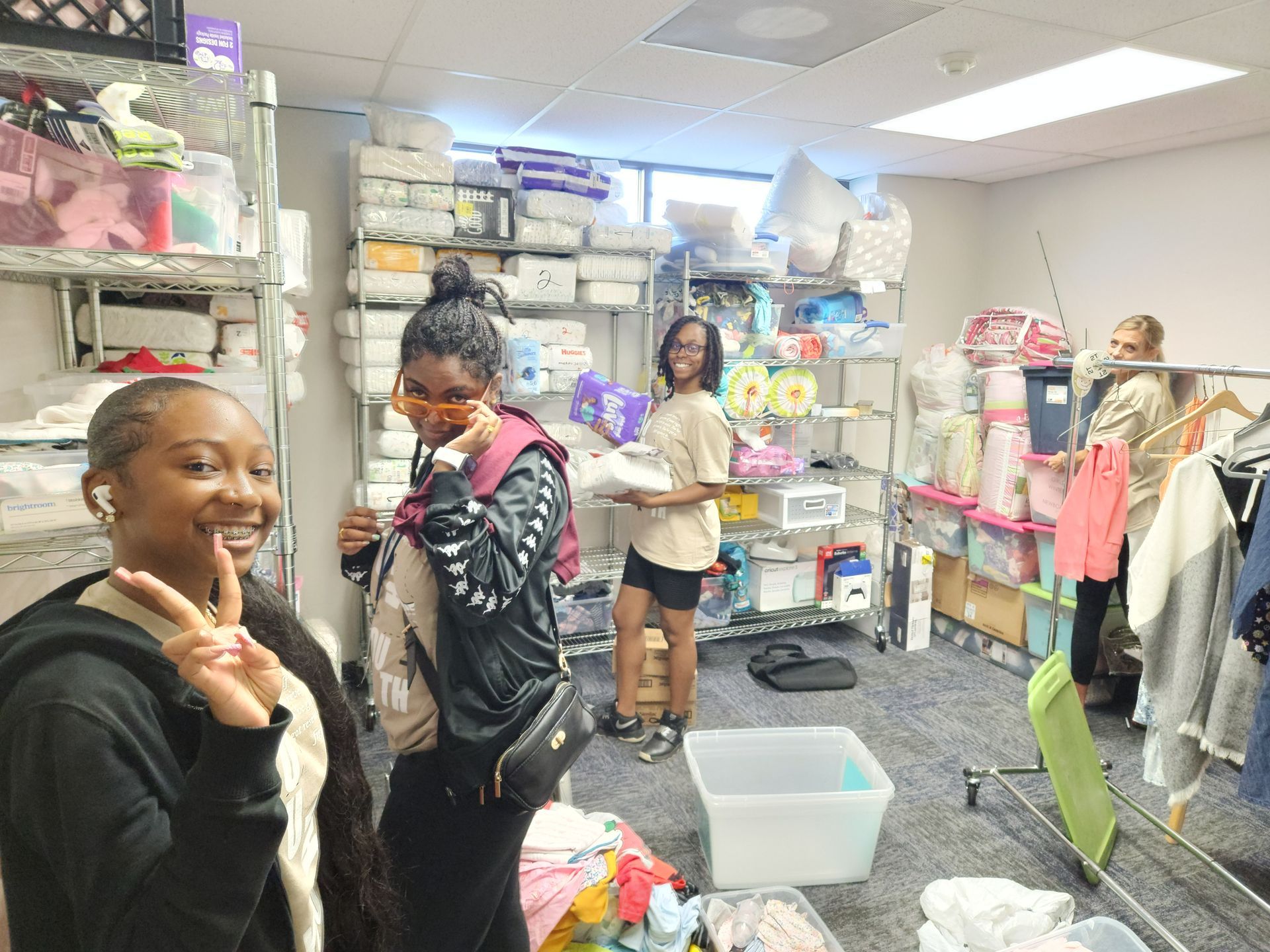 A group of girls are standing in a room filled with lots of clothes at an APRI charity drive