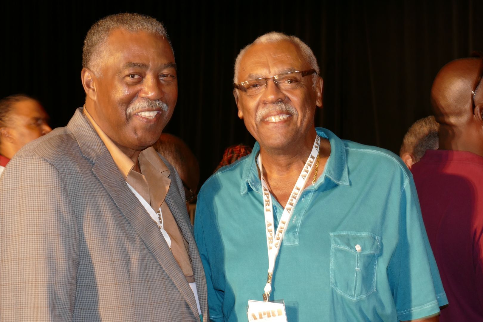 Two O.G.'s are posing for a picture and one of them has a lanyard around his neck that says ' APRI ' on it