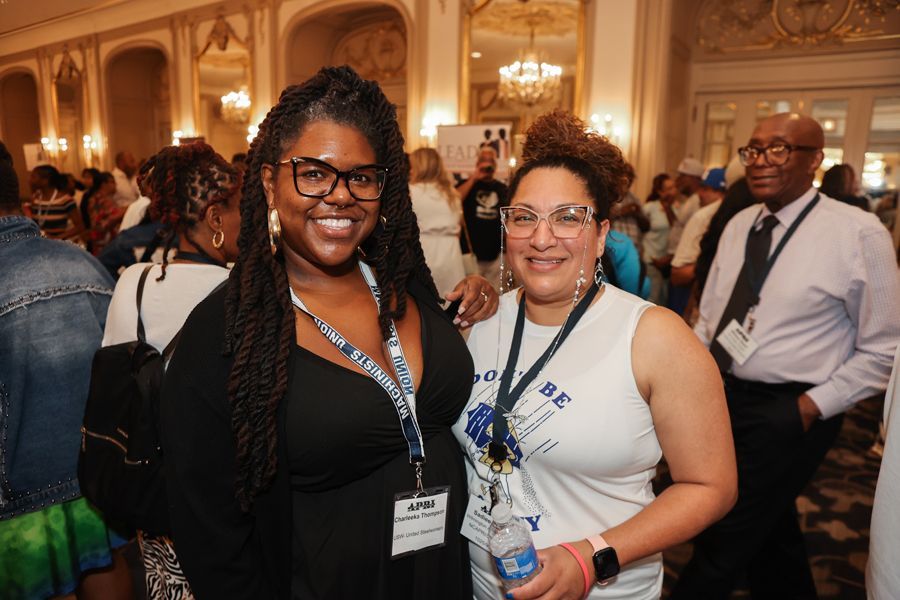 Two women smiling, posing together at an event in a well-lit room, likely at a conference or convention.
