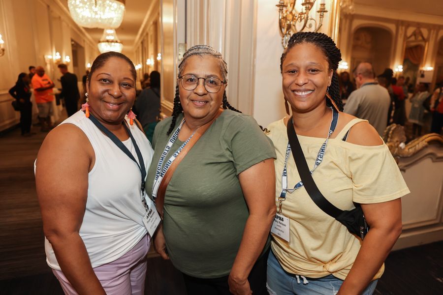 Three smiling women, indoors. Two wear light-colored tops, one has a green top. All have name tags.