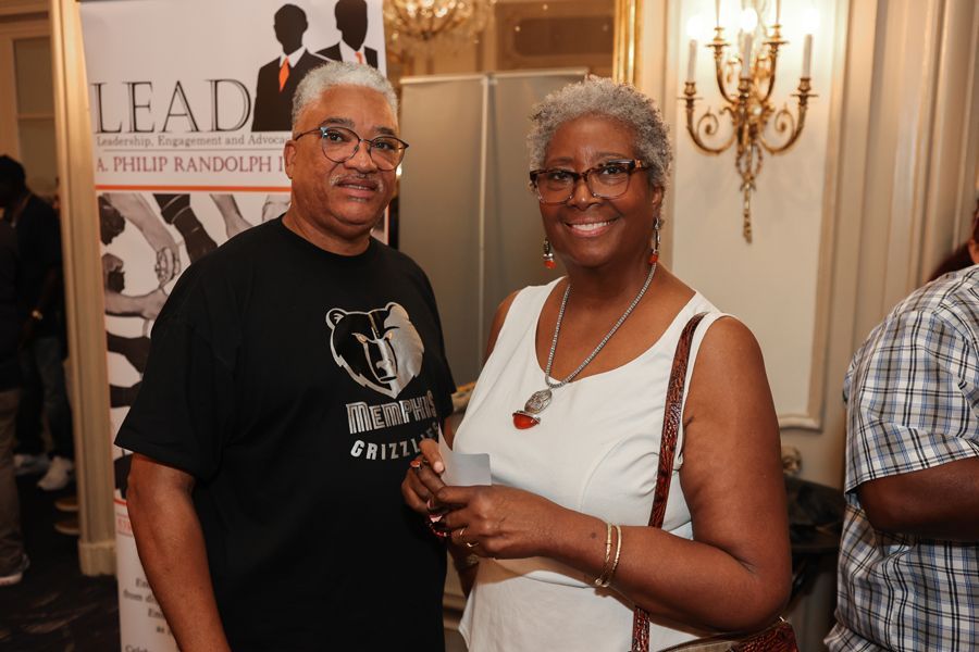 Man and woman smiling at an event, near a banner. They are in an ornate room with a chandelier.