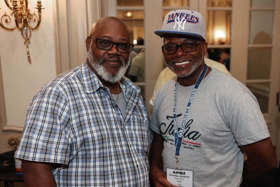 Two Black men pose; one in a plaid shirt, the other in a Yankees cap and a t-shirt, both smiling.