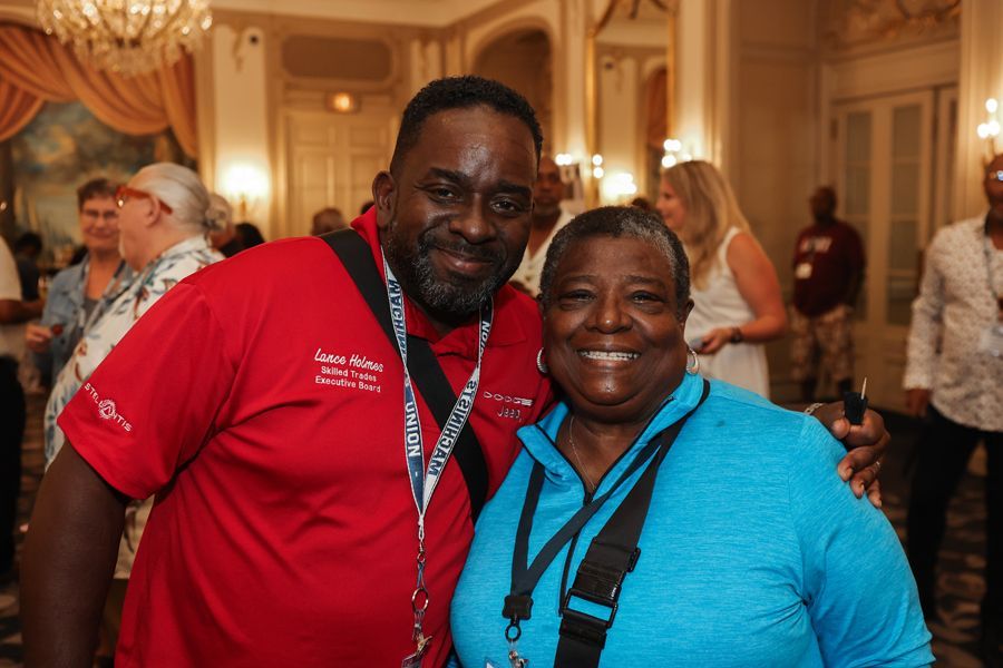 Two smiling people pose together at an event in a room. Man in red shirt, woman in blue shirt.