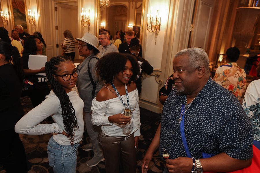 People socializing in a well-lit room with ornate decor. Three people in the foreground are conversing.