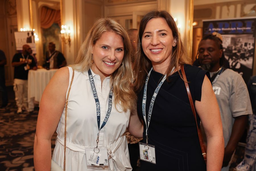 Two women smiling, arm in arm, at an event. White dress, black dress, lanyards, gold-trimmed room.