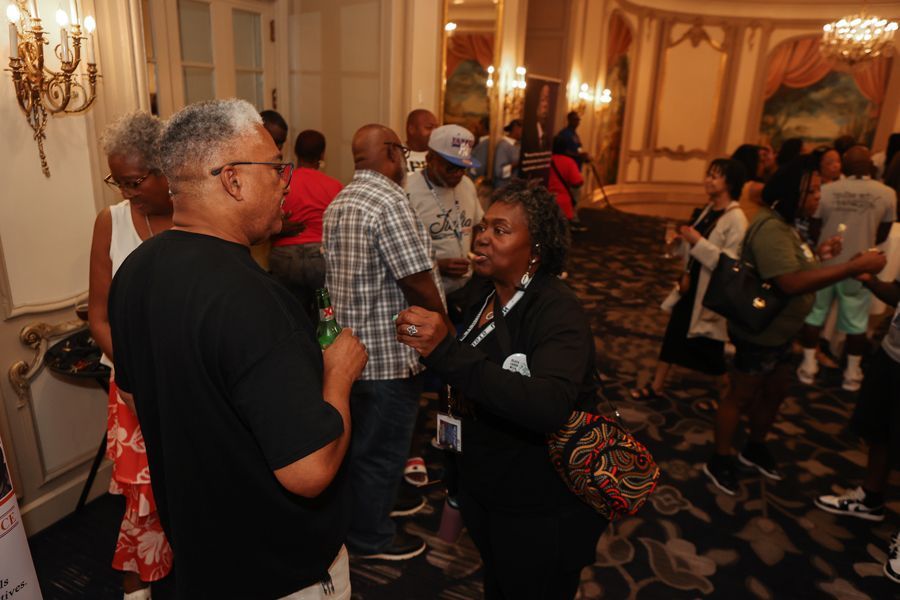 People conversing in a fancy room with ornate walls, chandeliers, and a mix of casual and formal attire.