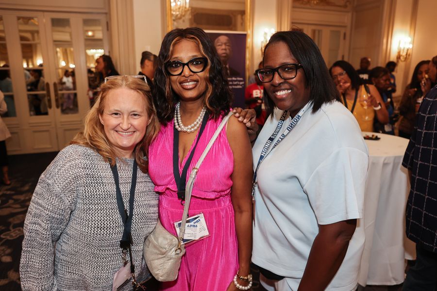 Three women pose together indoors; one in pink, one in blue, and one in a gray sweater; smiling at an event.