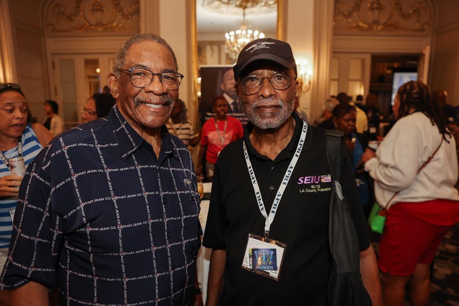 Two men smiling, posing together indoors, at an event. One in a checkered shirt, one in a black shirt and cap.