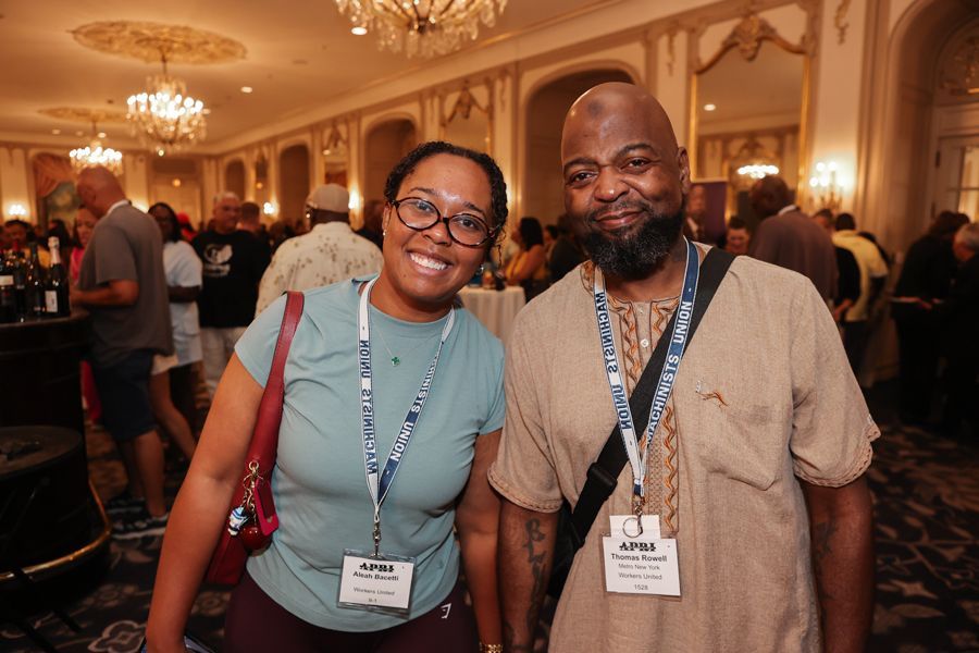 Two people smiling at an event, woman with glasses, man with beard, in a large ornate room.