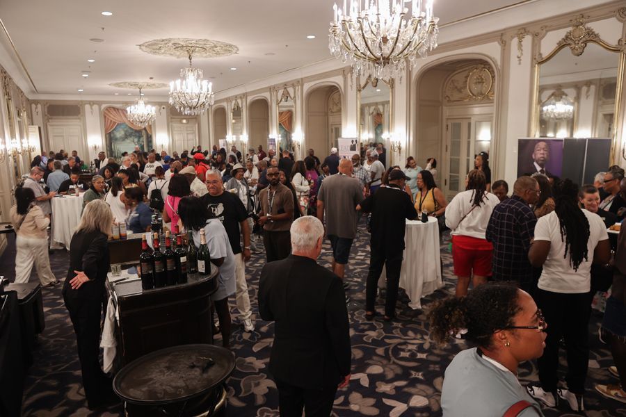 Large, crowded event in a decorated ballroom. People mingle, some near bar tables, chandeliers hang.