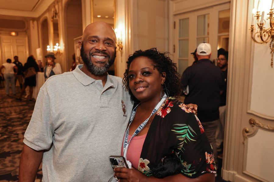 Smiling couple in an ornate hall; man with a gray shirt and a woman with a flowered top, at an event.