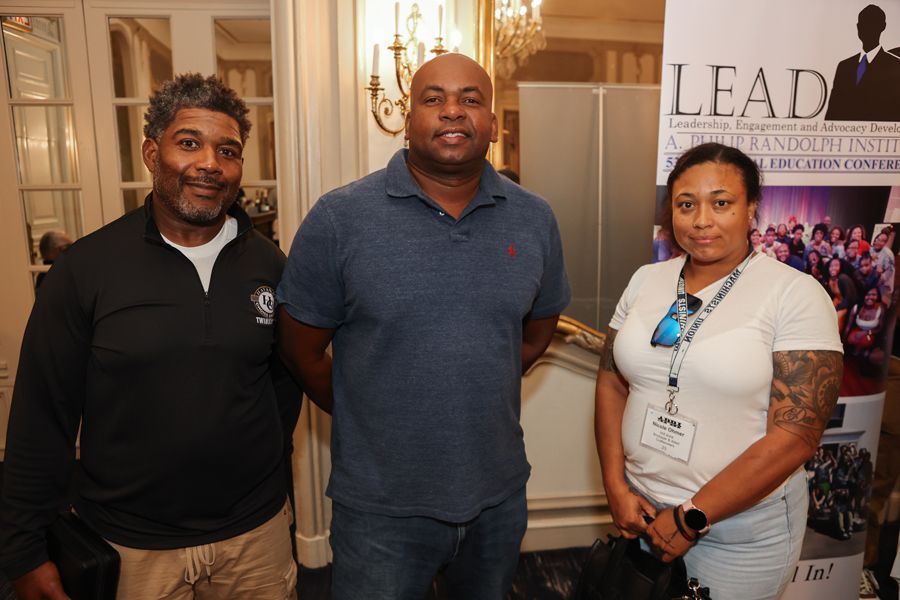Three people standing indoors at an event. Man in blue shirt flanked by a man and a woman, both with tattoos.