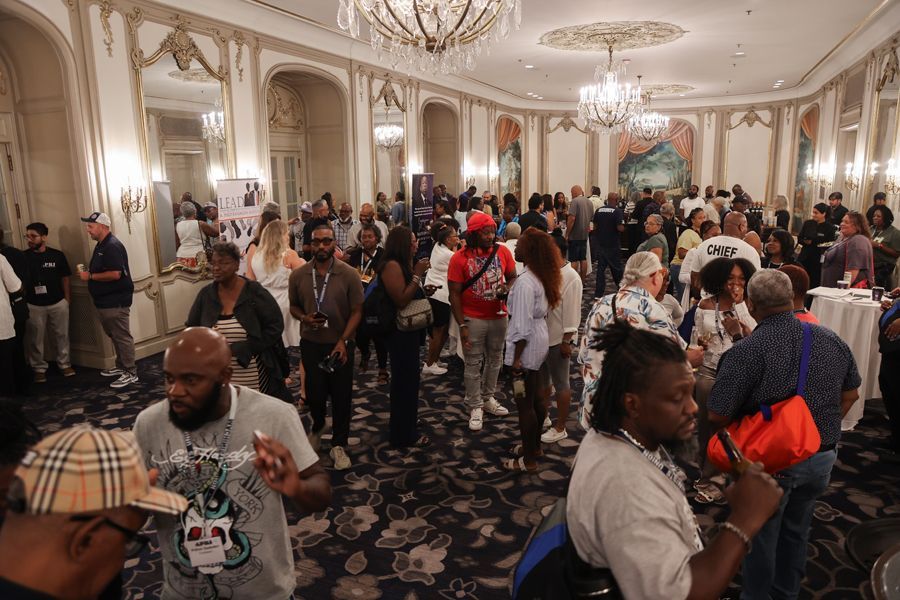 Large crowd in a ballroom. People socializing, wearing various outfits. Ornate decorations and chandeliers.