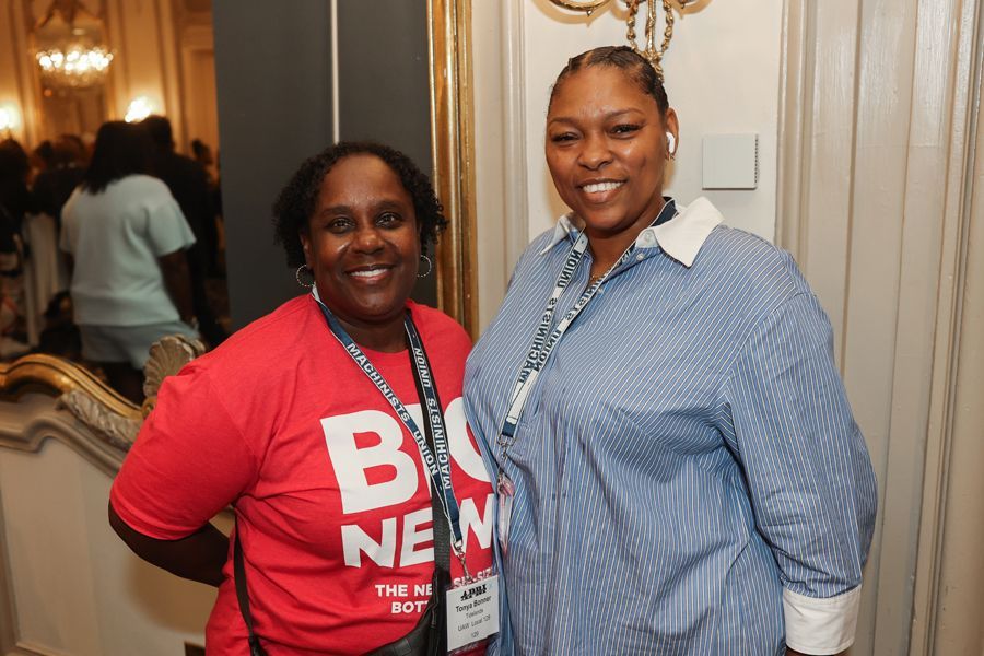 Two smiling women at an event. One in red shirt, one in blue striped shirt, both with lanyards.