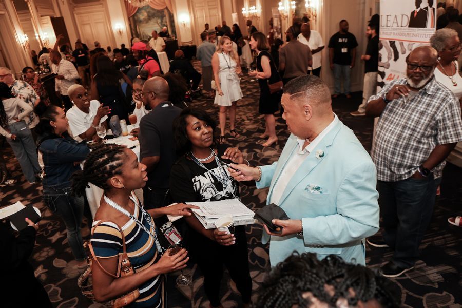 Conference attendees mingle, conversing. Man in light blue jacket speaks to women holding papers. Elegant room.