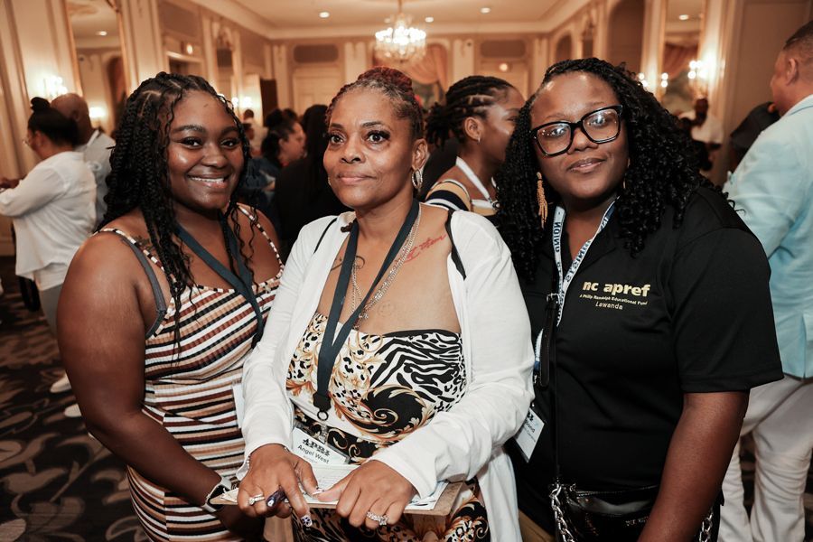 Three Black women at an event, smiling. One in a zebra print top, one in a striped dress, and one wearing glasses.