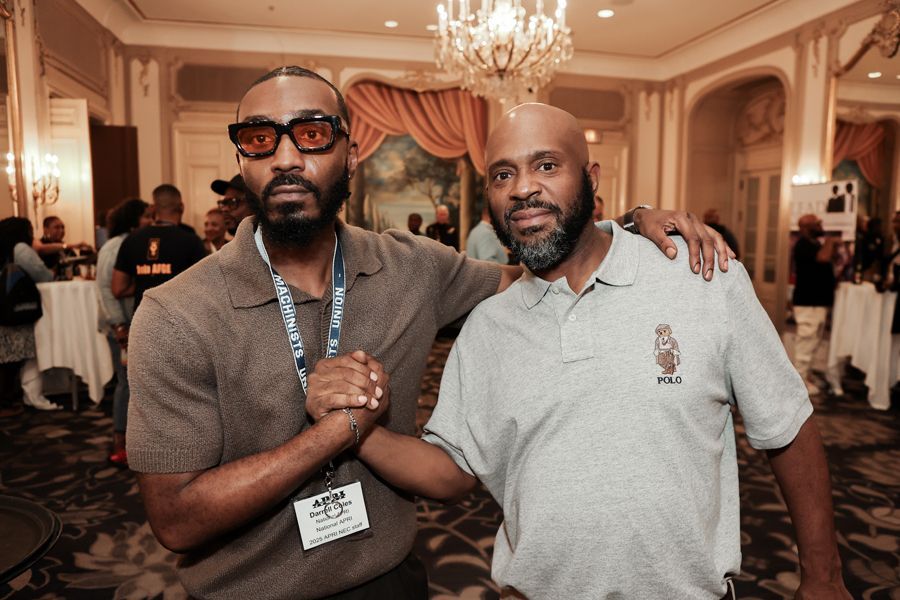 Two men pose, one with glasses, brown shirt, Al Brown in a polo shirt, arm around the other, holding hands, in a room.